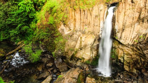 A serene waterfall cascading down rocky cliffs, surrounded by lush green foliage and a rocky riverbed below.