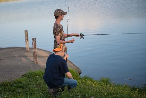 Two people fishing by a lake, one standing and casting a line, the other sitting on the grass nearby.