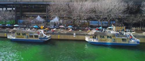 Two boats docked at a waterfront with bare trees and tents in the background, reflecting in green water.