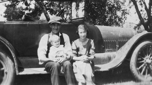 A vintage black-and-white photo of a man, woman, and baby sitting on the fender of an old car.