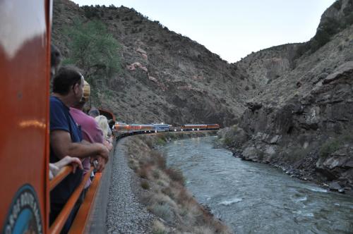 Passengers on a train admire the scenic river and rocky landscape as the train winds through a canyon.