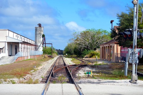 A view of railway tracks splitting, with industrial buildings and trees in the background under a blue sky.