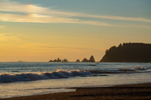 Sunset over a calm ocean with gentle waves, rocky formations in the distance, and a forested shoreline.