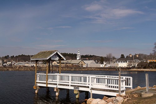 A white dock extends into a calm lake, with a small town and church steeple visible in the background under a blue sky.