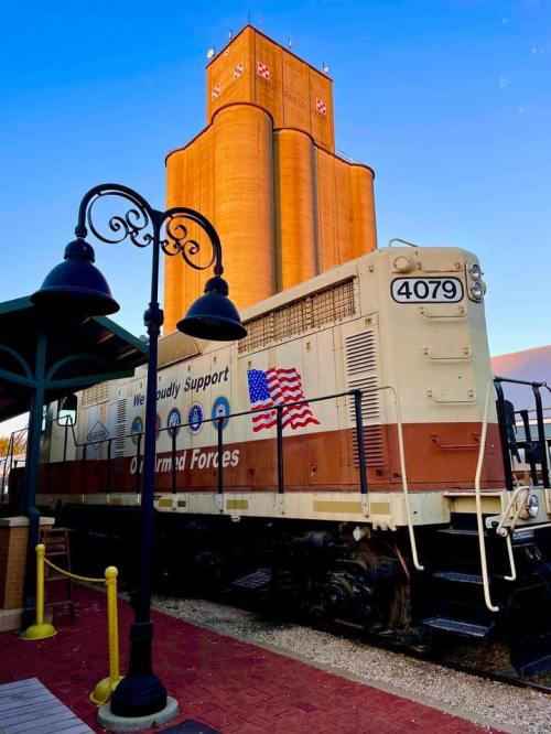 A vintage train engine with a patriotic design parked near a tall, orange building against a clear blue sky.