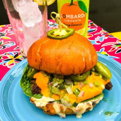A colorful burger with a bright orange bun, lettuce, jalapeños, and cheese, served with a drink and ice.