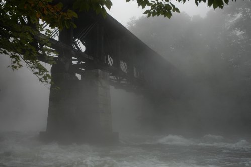 A foggy scene featuring a weathered bridge over a rushing river, surrounded by misty trees.