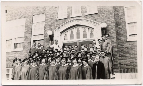 A large group of graduates in caps and gowns poses in front of a brick building, smiling for the camera.