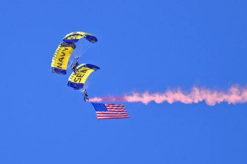 Two parachutists descend with a large American flag, trailing pink smoke against a clear blue sky.