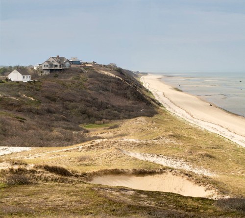 A coastal landscape featuring sandy dunes, a beach, and houses on a cliff overlooking the ocean under a cloudy sky.