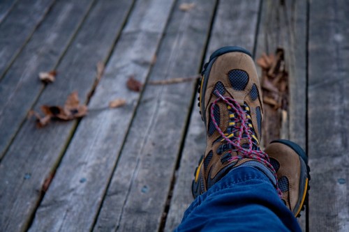 A close-up of a person's feet in hiking boots resting on a wooden deck, with leaves scattered nearby.