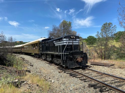A black and silver train with a passenger car travels along a scenic track under a blue sky with wispy clouds.