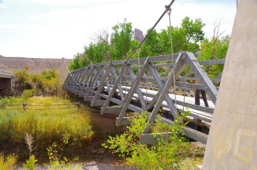 A wooden suspension bridge spans a small area, surrounded by greenery and rocky terrain under a clear sky.