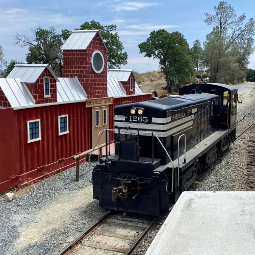 A vintage black and silver train near a red barn-style building on a gravel track, surrounded by trees.