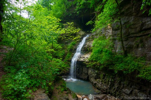 A serene waterfall cascades over rocks, surrounded by lush green foliage and trees in a tranquil forest setting.