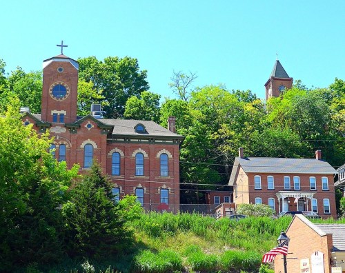 Historic brick buildings with a clock tower and cross, surrounded by lush greenery under a clear blue sky.