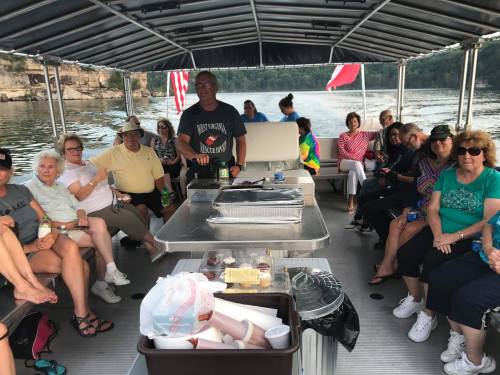 A group of people enjoying a boat ride, seated around a table with food, on a scenic lake.