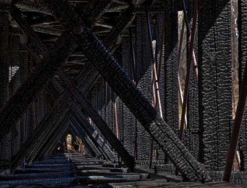 Charred wooden beams create a geometric pattern under a bridge, with light filtering through the gaps.