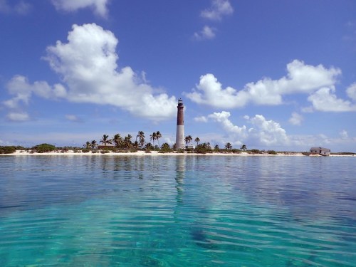 A serene coastal scene featuring a lighthouse on an island, surrounded by clear turquoise waters and fluffy clouds.