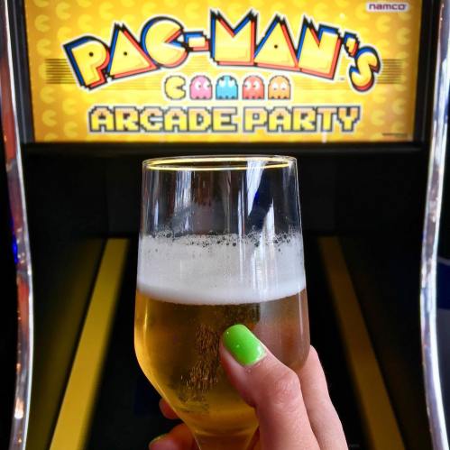 A hand holding a glass of beer in front of a Pac-Man arcade game screen displaying "Pac-Man's Arcade Party."
