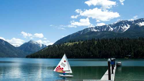 A sailboat with a colorful sail glides on a serene lake, surrounded by mountains and a clear blue sky.