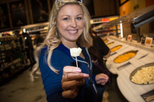 A smiling woman holds a piece of cheese on a toothpick in a food store, with various cheeses displayed in the background.