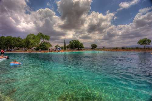 A serene swimming area with clear blue water, surrounded by trees and a cloudy sky, with people enjoying the space.