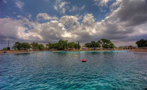 A serene swimming area with clear water, surrounded by trees and cloudy skies, with people enjoying the space.