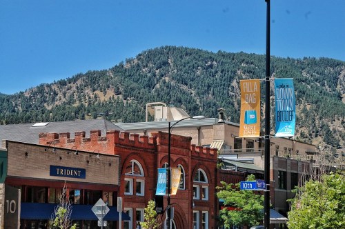 A vibrant street scene with brick buildings, banners, and a backdrop of green mountains under a clear blue sky.