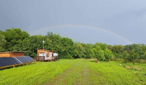 A lush green field with a wooden cabin, solar panels, and a wind turbine under a cloudy sky with a rainbow.
