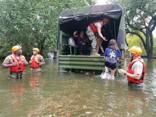 Rescue team assists people from a military truck in a flooded area, with water surrounding them and trees in the background.