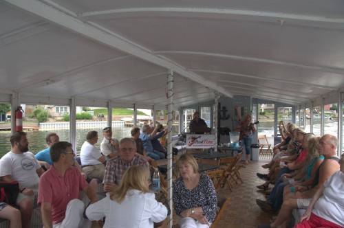A group of people seated indoors on a boat, enjoying a gathering with music and a view of the water.