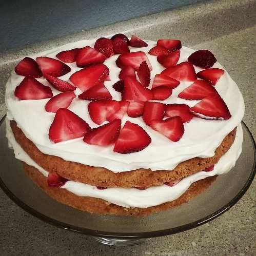 A layered cake topped with fresh strawberries and whipped cream, displayed on a glass cake stand.