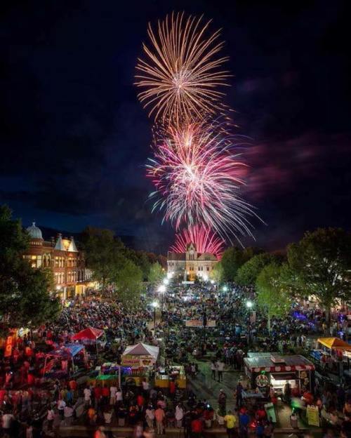 A vibrant fireworks display lights up the night sky over a crowded outdoor event, with trees and buildings in the background.