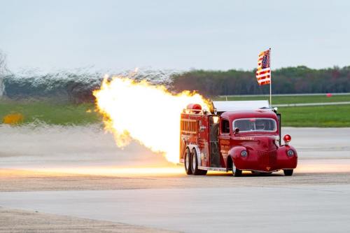 A vintage red fire truck with flames shooting out the back, driving on an airstrip with an American flag.