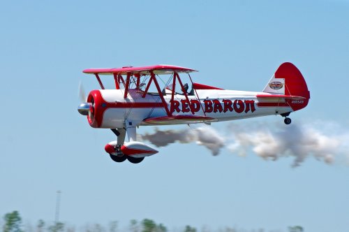 A red and white biplane labeled "Red Baron" performs a stunt, trailing smoke against a clear blue sky.