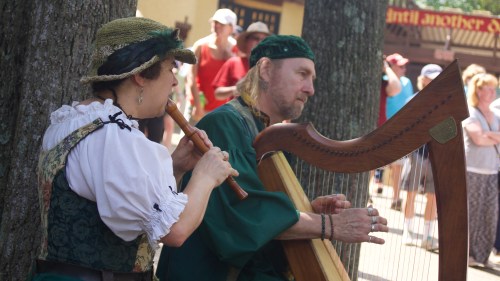 Two musicians perform outdoors, one playing a flute and the other a harp, with a crowd in the background.