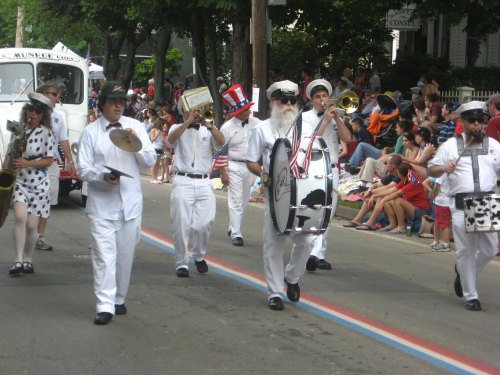 A marching band in white uniforms parades down a street, with spectators lining the sides, celebrating a festive event.