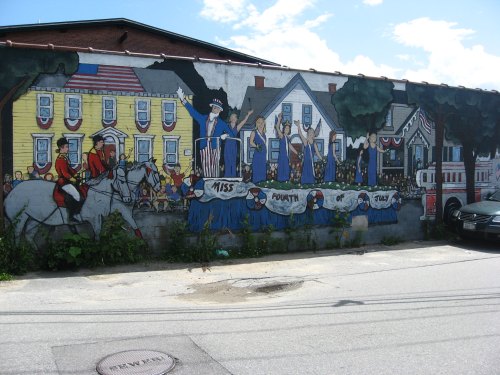 A colorful mural depicting a Fourth of July parade with people, horses, and festive decorations on a building wall.