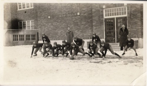 A vintage black-and-white photo of a football team in snowy conditions, posing in front of a brick school building.