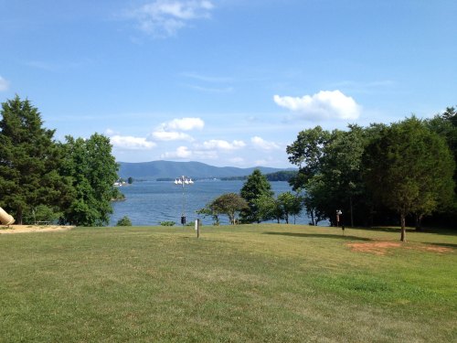A serene lake view with green grass, trees, and distant mountains under a clear blue sky.