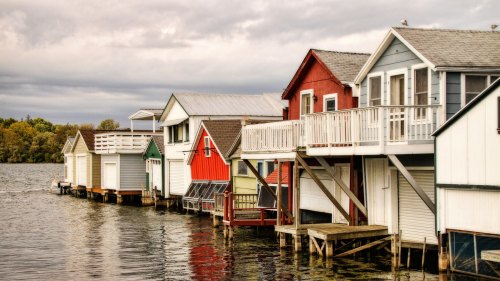 Colorful boathouses line a calm lake, reflecting in the water under a cloudy sky.