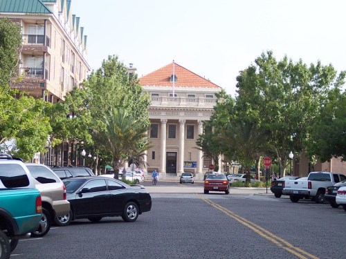 A view of a historic building with a red-tiled roof, surrounded by trees and parked cars on a sunny day.