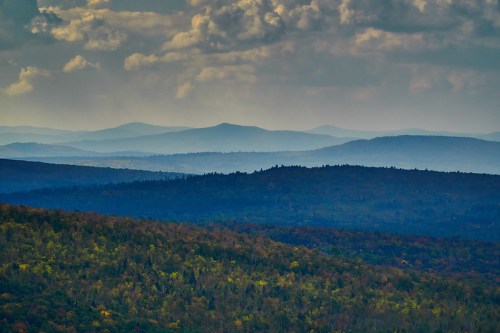 A panoramic view of layered mountains under a cloudy sky, with lush green forests in the foreground.
