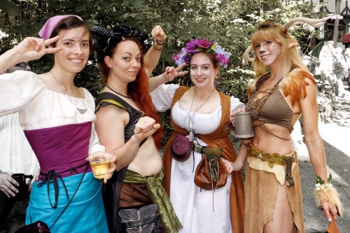 Four women in colorful costumes pose together outdoors, smiling and making playful gestures.