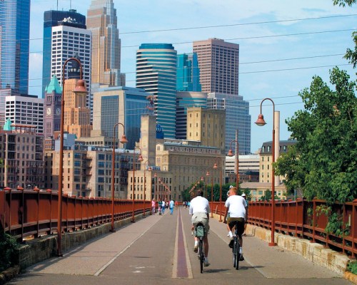 Two cyclists ride on a bridge with a city skyline in the background, featuring tall buildings and clear blue skies.