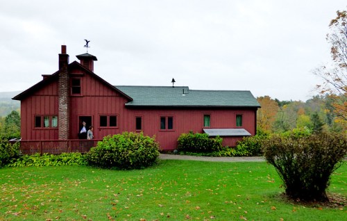 A red barn-style house with a stone chimney, surrounded by green grass and trees, on a cloudy day.