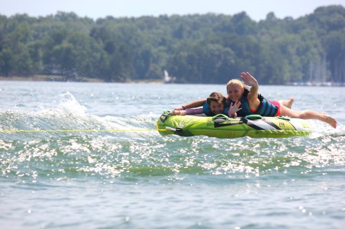 Two children enjoy tubing on a lake, smiling and waving as they glide over the water.