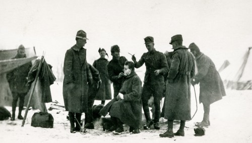 Soldiers in winter attire gather around a man getting a haircut in a snowy camp setting.