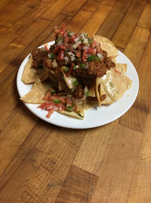 A plate of nachos topped with fried chicken, pico de gallo, and fresh herbs on a wooden table.